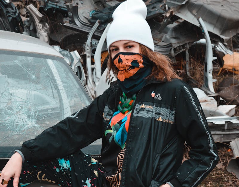 A young woman in a beanie and mask poses by a broken car in a junkyard.