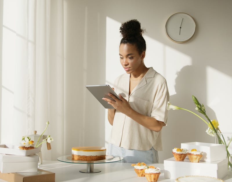 A woman using a tablet in a bright and modern kitchen setting, surrounded by cakes and flowers, depicting a lifestyle scene.
